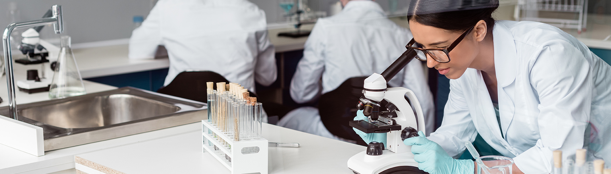 Kvinna sitter i ett lab och tittar i ett mikroskop. I bakgrunden syns hennes kollegor.