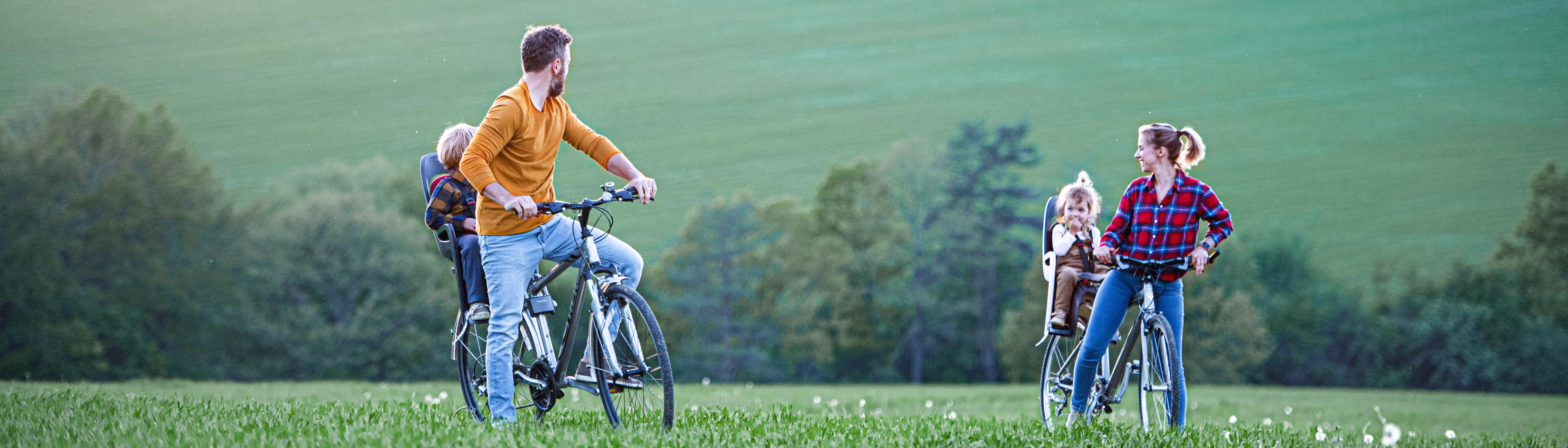 Familj cyklar en sommardag. En man och en kvinna cyklar på en stor grön äng. Båda har ett barn bakom sig på cyklarna.