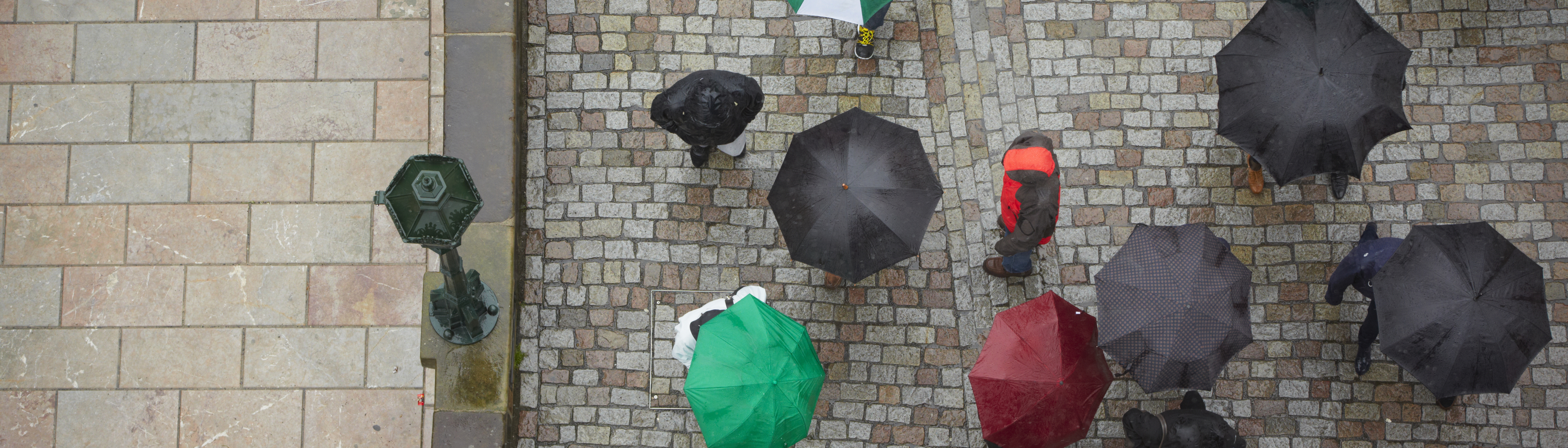 Bild tagen uppifrån på en gata med kullersten där det går folk med paraplyn.